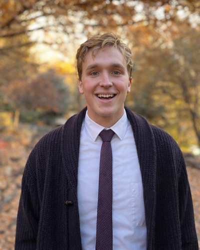 Jackson Pond wearing a shirt, tie, and sweater smiling outside with fall foliage