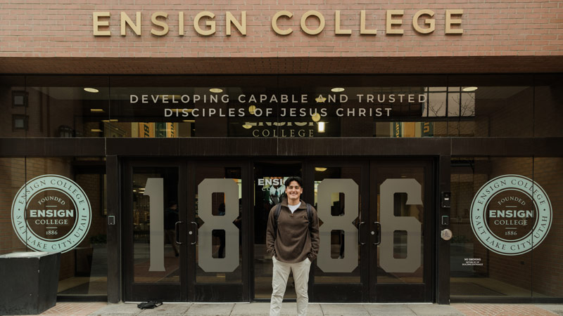 A male Ensign College student standing in front of the main Ensign College building